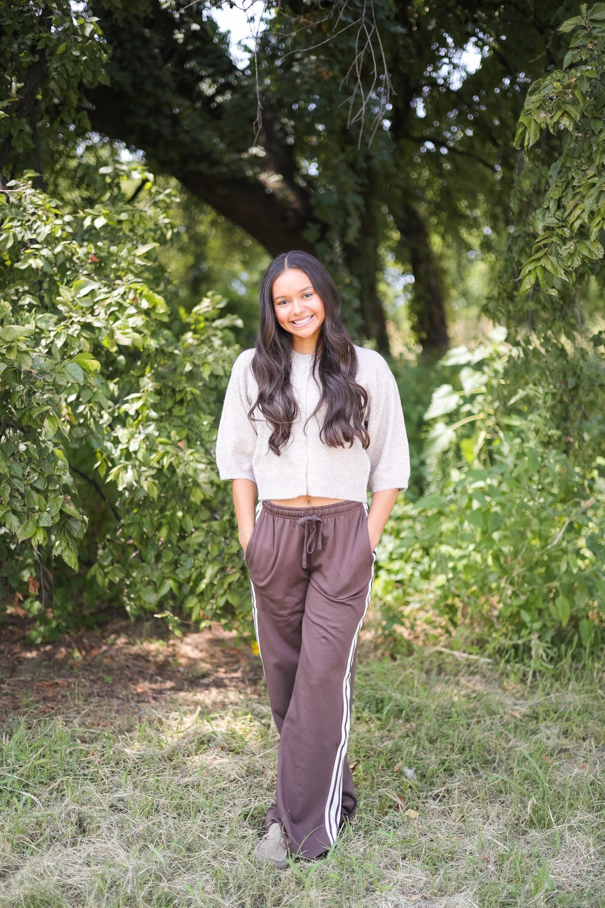 Woman wearing a beige cropped sweater and brown side-stripe joggers in a wooded outdoor setting.