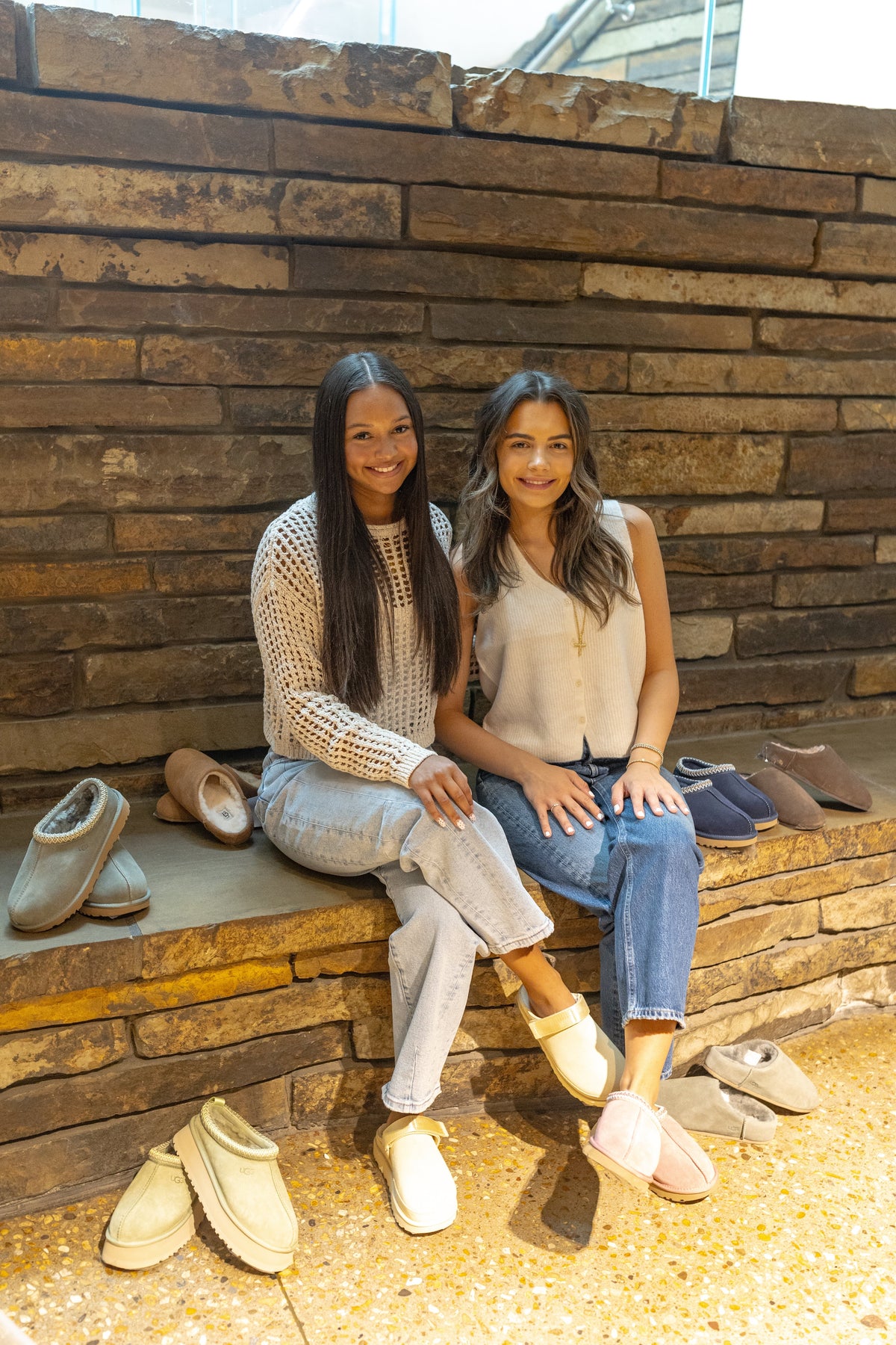 Two women sitting on a stone ledge surrounded by various cozy fall boots and slippers.