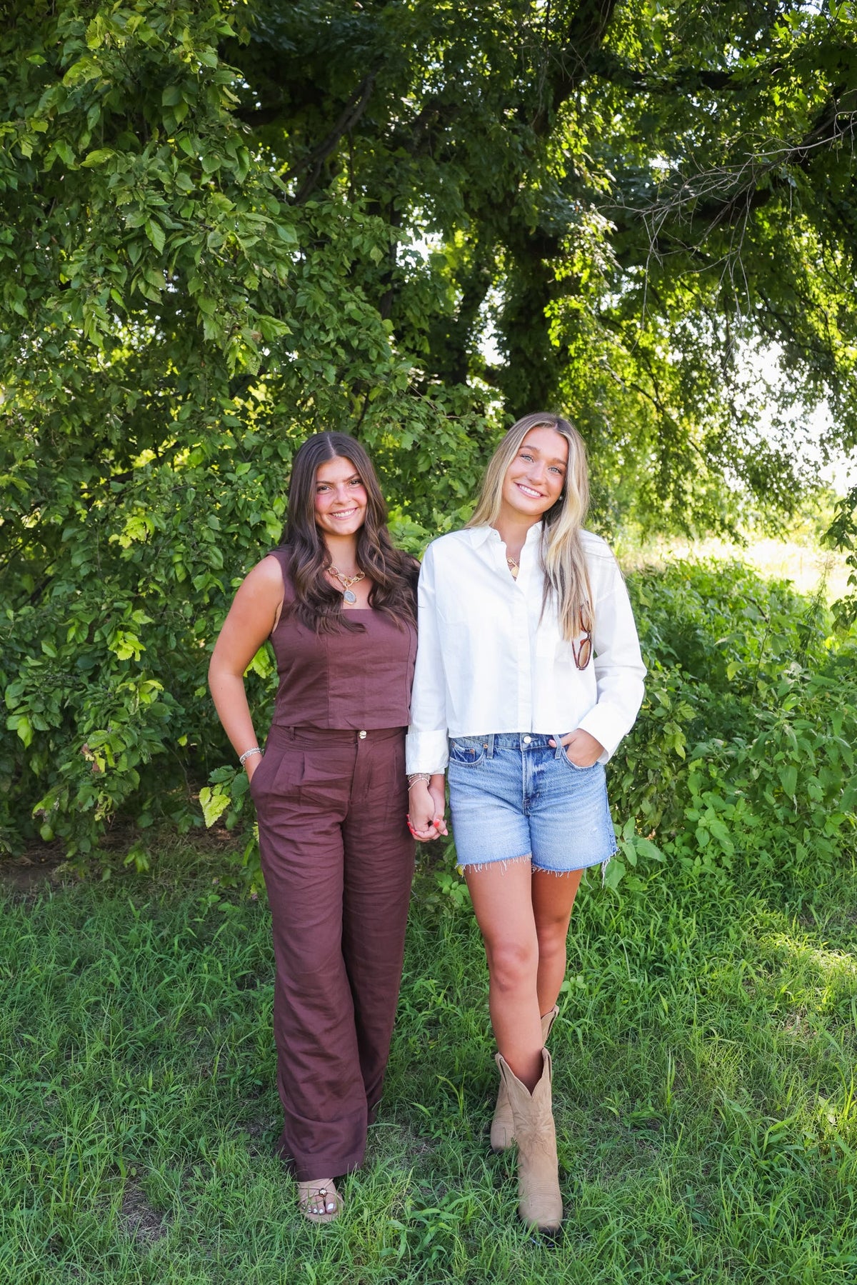 Two women posing outdoors in a brown jumpsuit and a white button-down shirt with denim shorts.