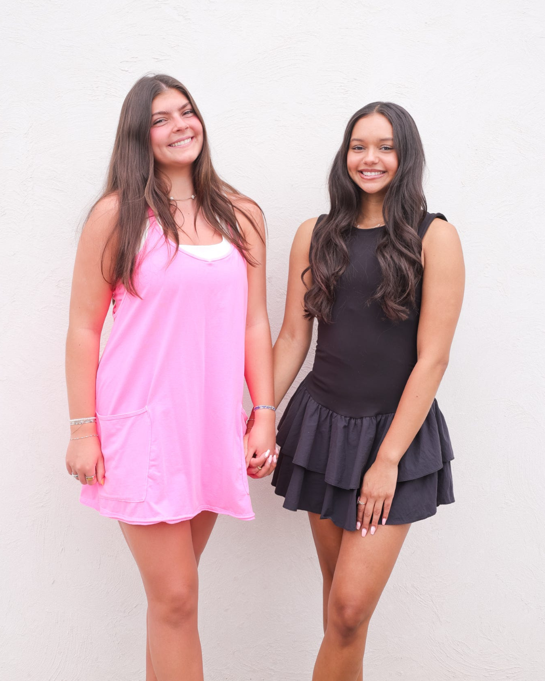 Two women holding hands, one in a pink sleeveless dress and one in a black ruffled mini dress.