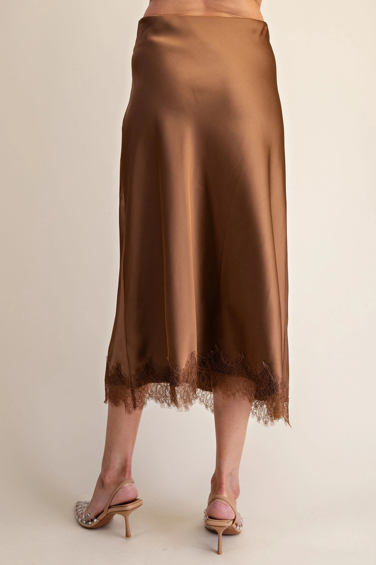 A brown satin midi skirt with a lace hem, shown on a model from the back against a light beige studio background.