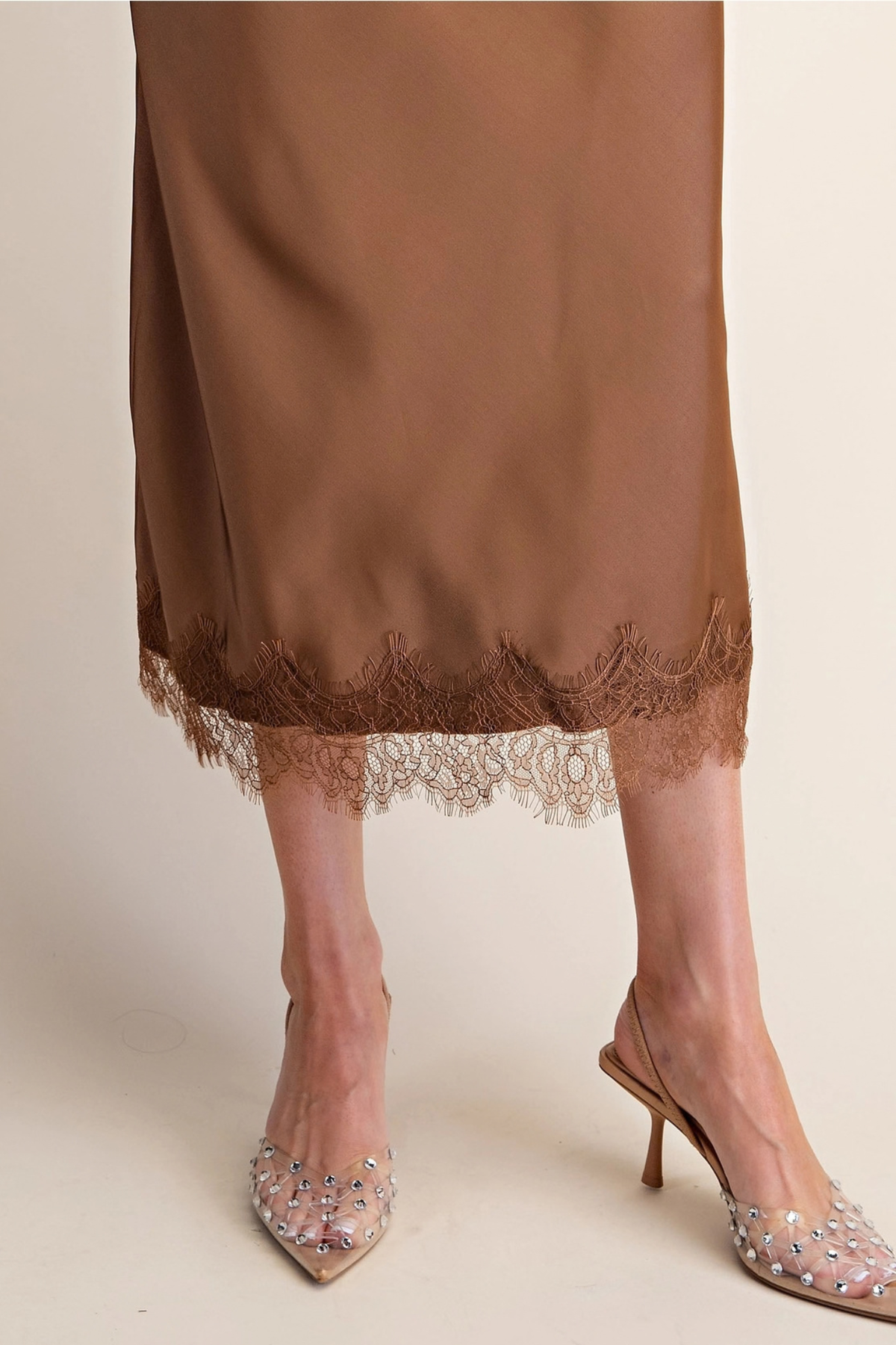 A close-up of a person wearing a brown satin slip skirt with a lace hem, paired with embellished nude slingback heels, against a plain beige studio background.