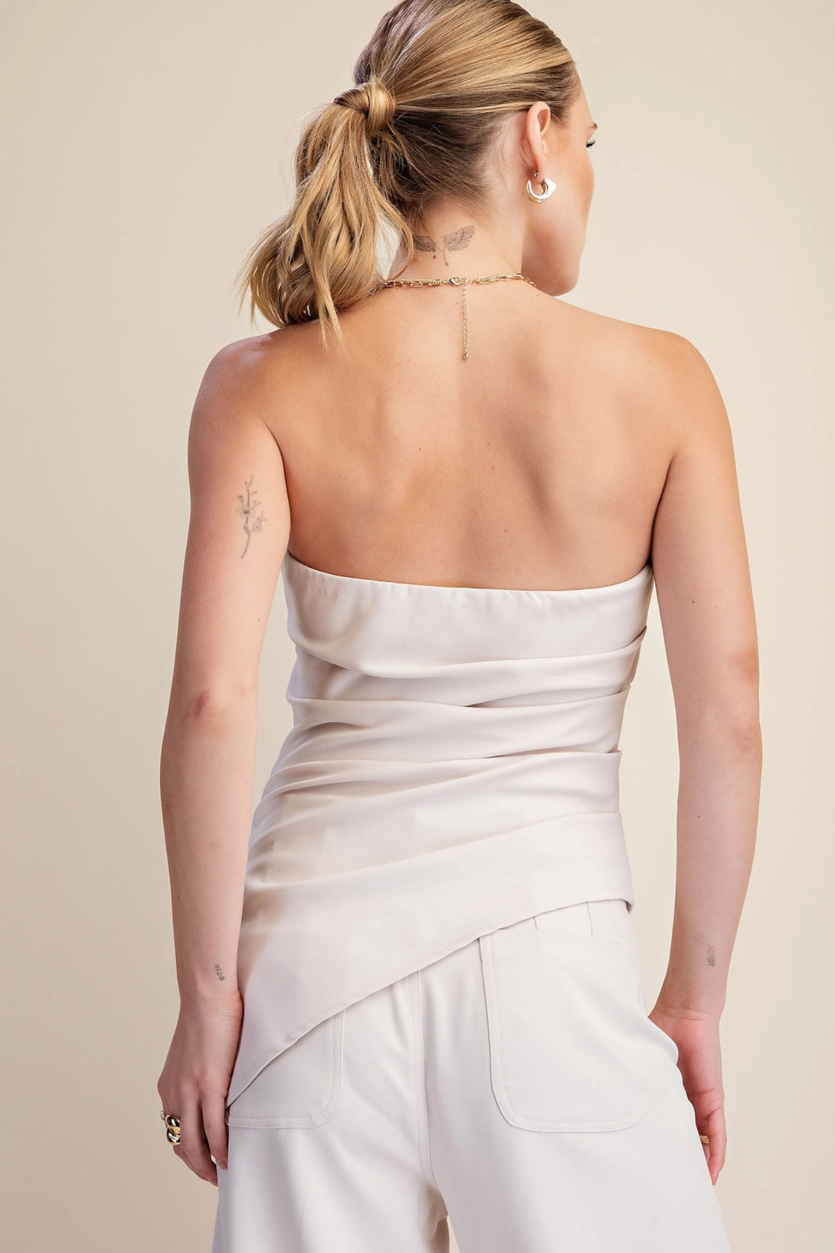 A woman shown from the back wearing a strapless off-white top with a draped, gathered waist and matching off-white pants against a plain beige studio background.