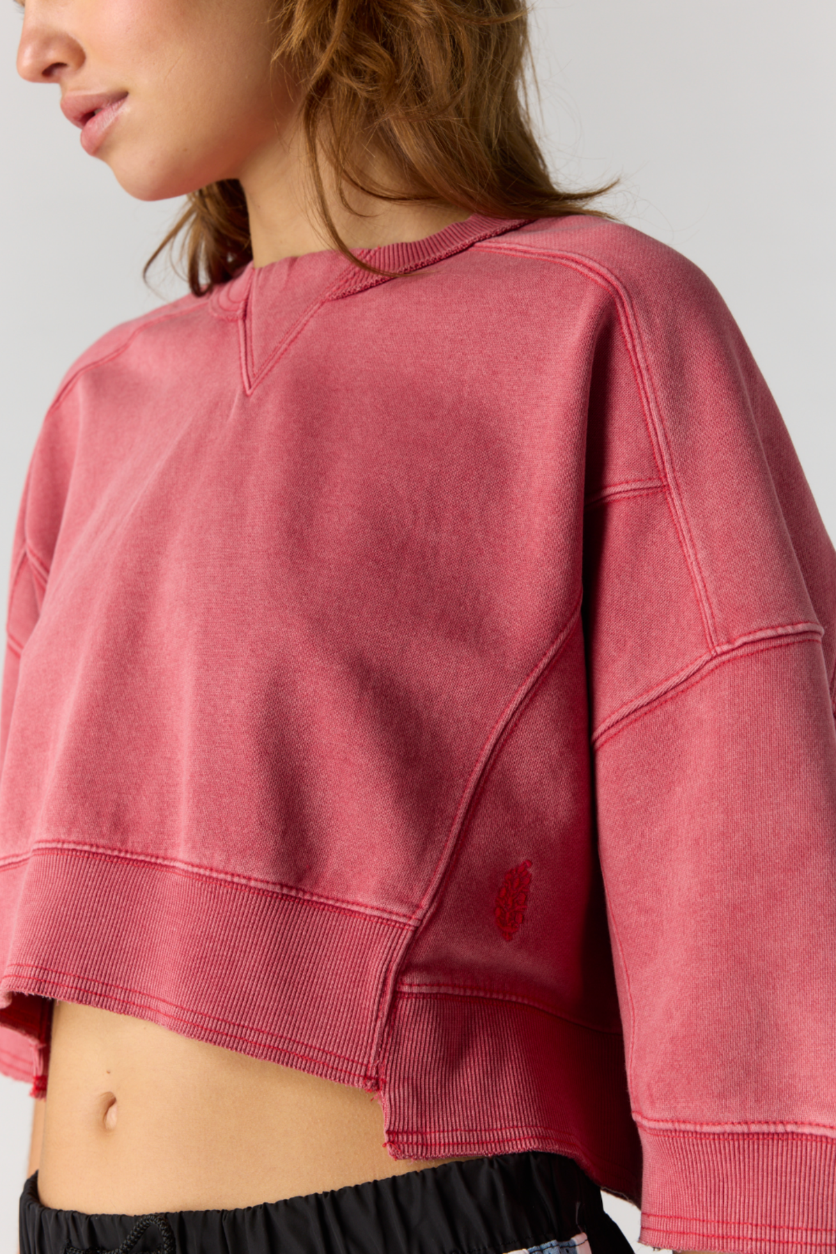 A close-up of a model wearing a cropped, washed red sweatshirt with long sleeves and ribbed trim, paired with black bottoms against a light gray background.