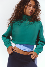 A model with curly brown hair wearing a green cropped mock-neck sweatshirt, a light blue ribbed tank top, and dark navy pants, posing against a white studio background.