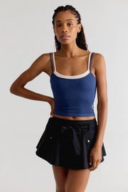 A woman modeling a blue ribbed tank top with white trim and a black pleated mini skirt against a light gray studio background.