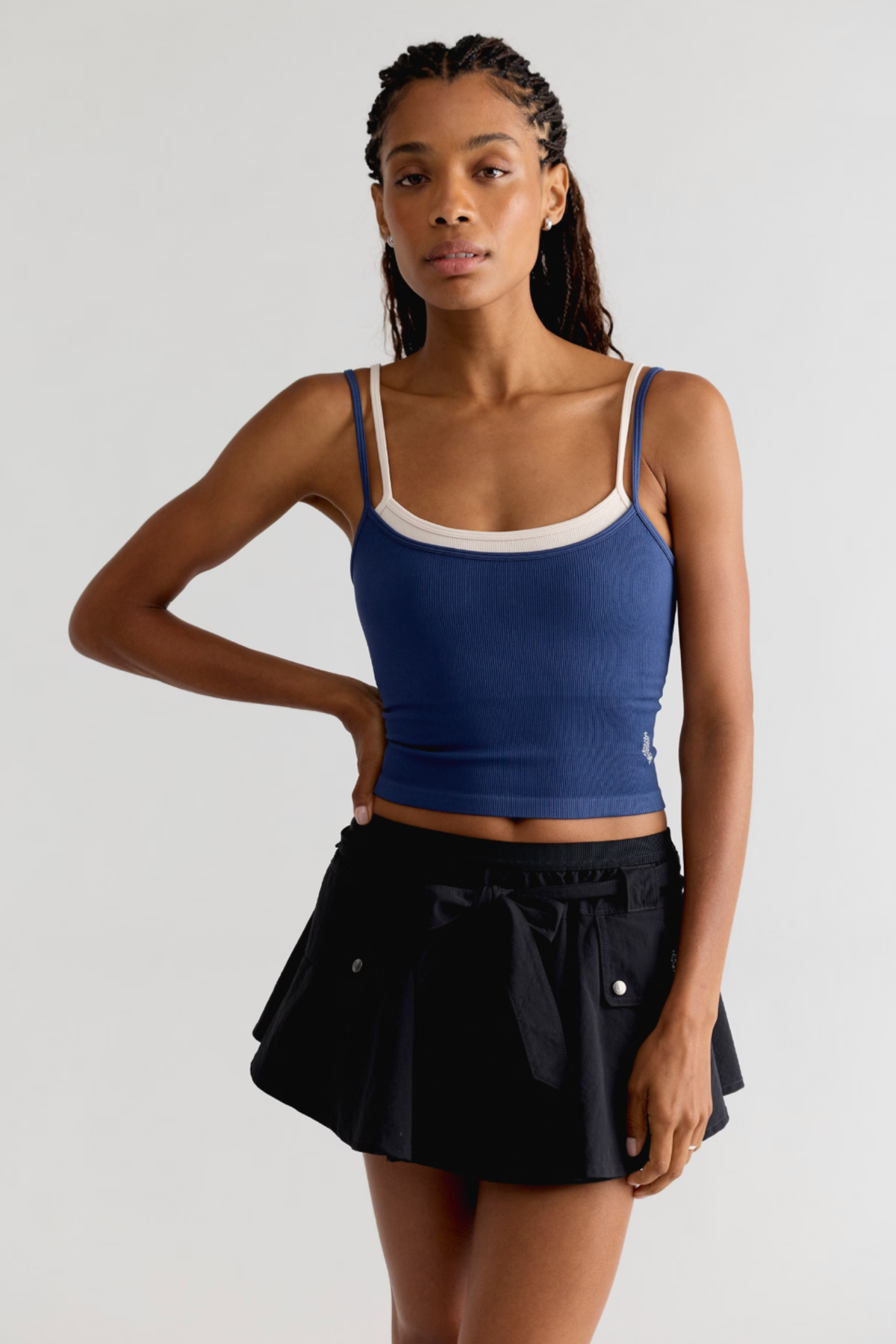 A woman modeling a blue ribbed tank top with white trim and a black pleated mini skirt against a light gray studio background.