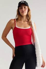 A woman modeling a bright red ribbed tank top with a white inner band, paired with black leggings and a black baseball cap, while carrying a white puffer bag over her shoulder against a light gray studio background.
