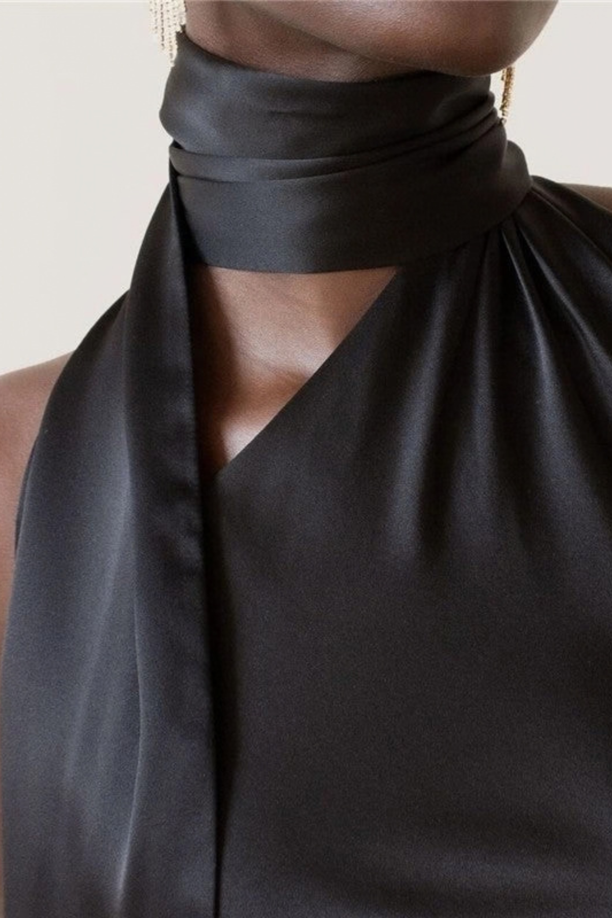 Close-up of a model wearing a black satin halter top with a draped high neck and side cutout, paired with gold drop earrings against a light beige background.