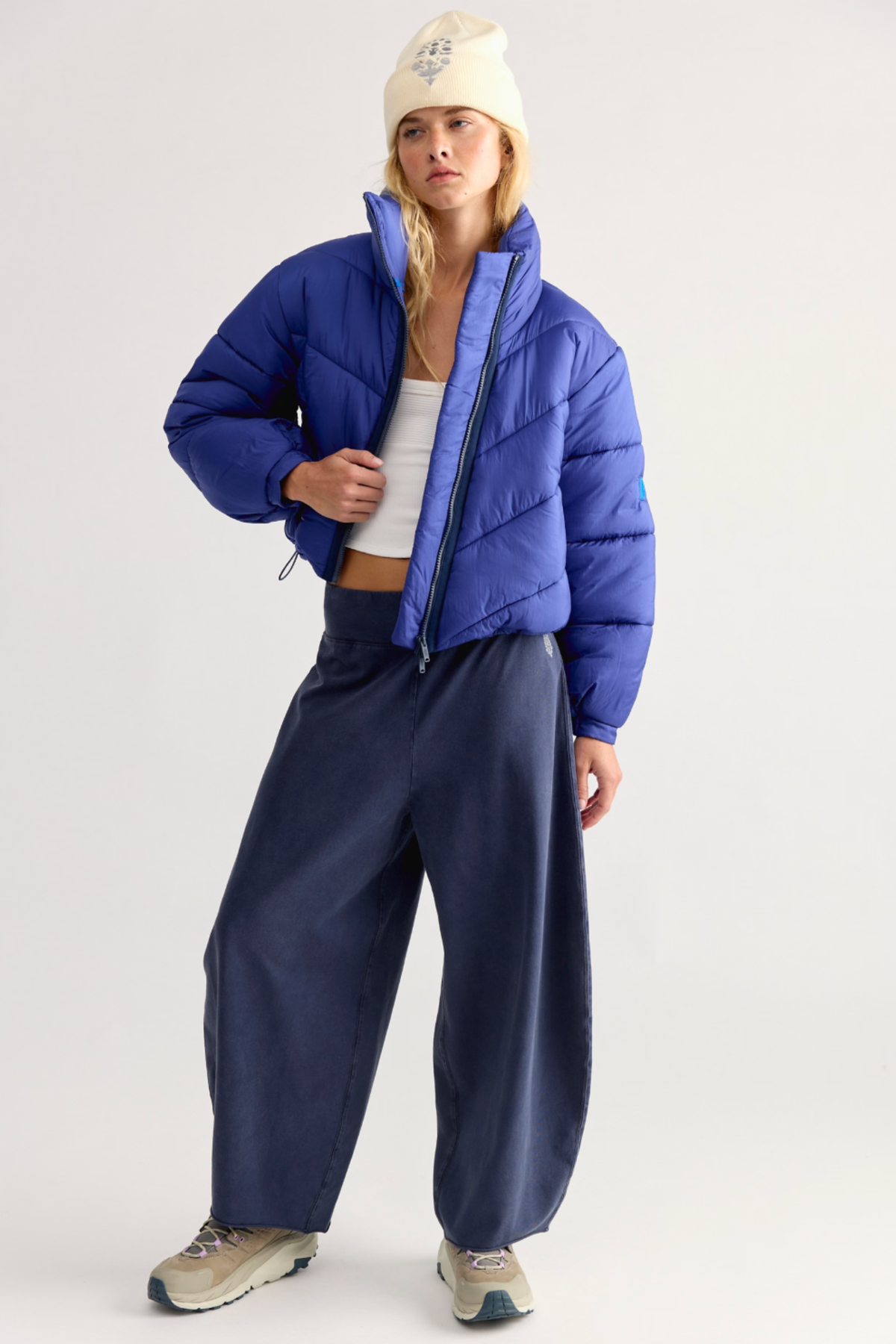 A model wearing a bright blue cropped puffer jacket, navy wide-leg pants, a cream beanie, and beige sneakers, standing against a light gray studio background.