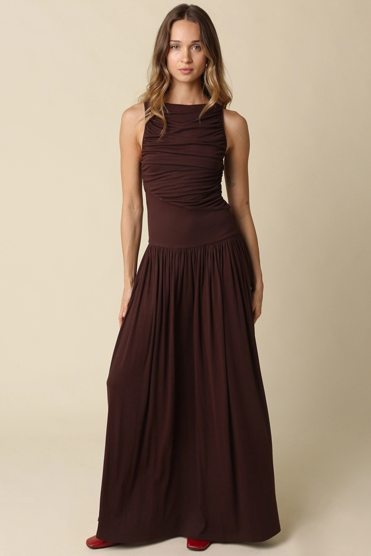 A woman modeling a sleeveless, high-neck, floor-length dark brown dress against a plain beige studio background.