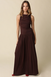 A woman modeling a sleeveless, high-neck, floor-length dark brown dress against a plain beige studio background.