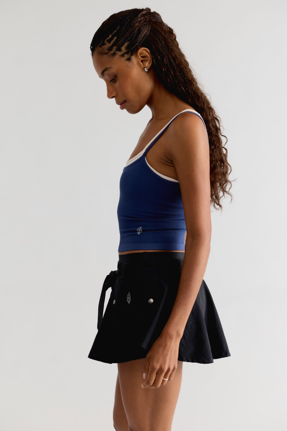A woman wearing a blue ribbed crop tank top with white trim and a black pleated mini skirt with a tie waist, photographed against a plain light gray studio background.