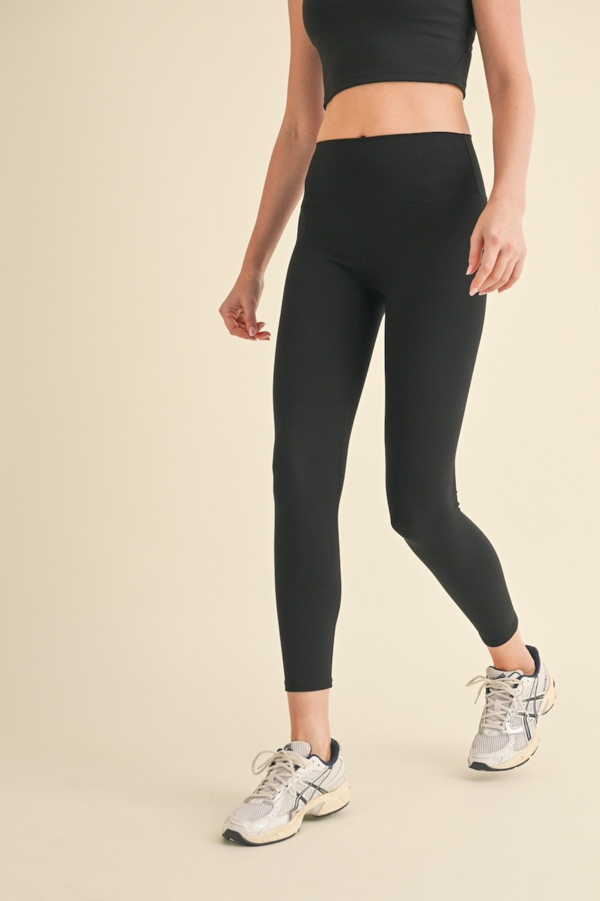 A model wearing black high-waisted athletic leggings and a matching black crop top, paired with white and gray sneakers, against a solid beige background.