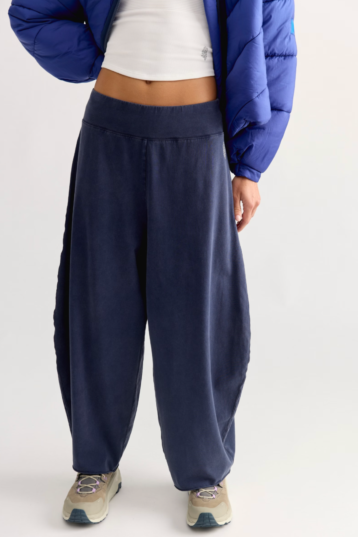A person wearing loose navy blue sweatpants with a white cropped top and a blue puffer jacket, standing against a light gray studio background.