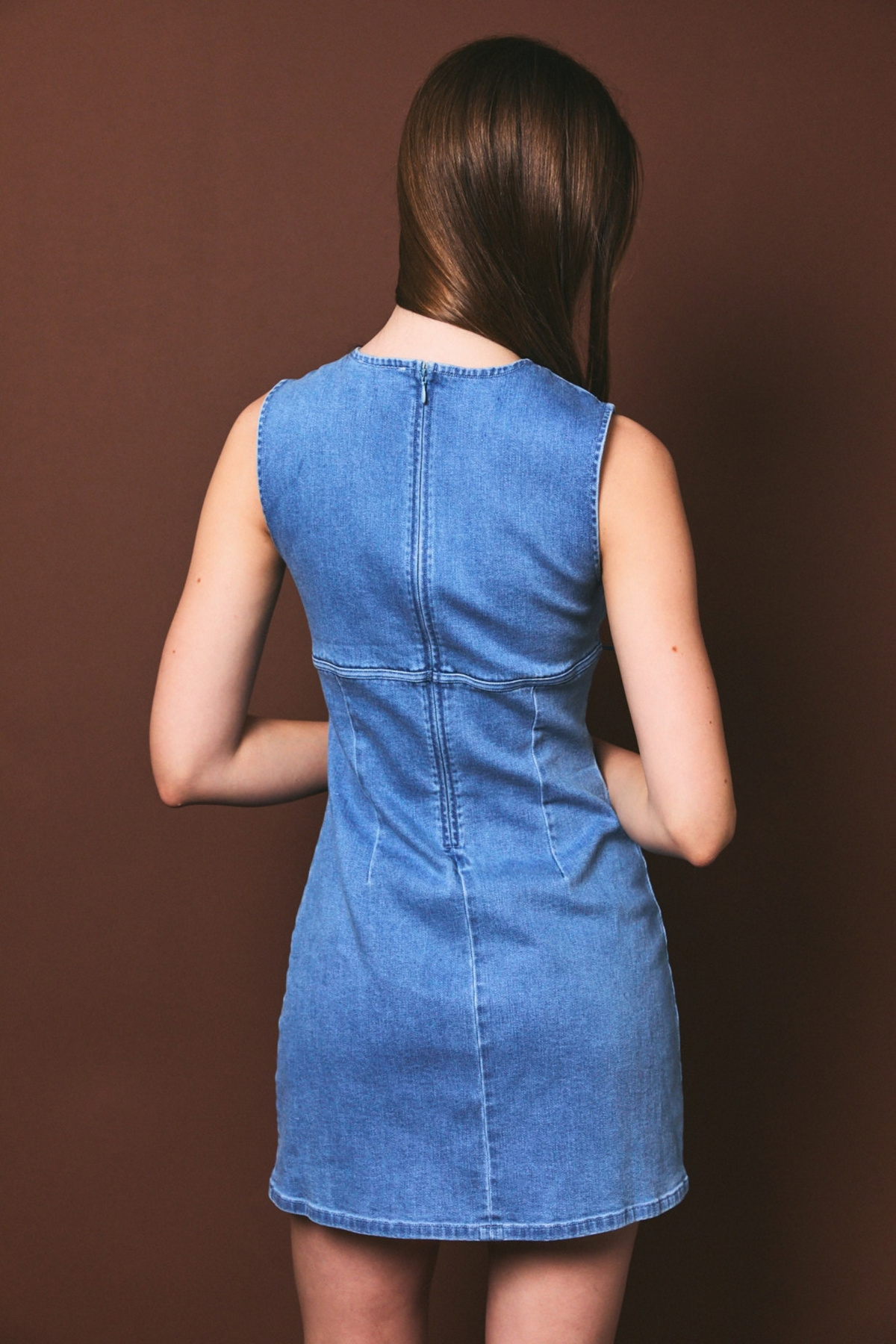A woman viewed from behind wearing a sleeveless blue denim dress against a solid dark brown studio background.