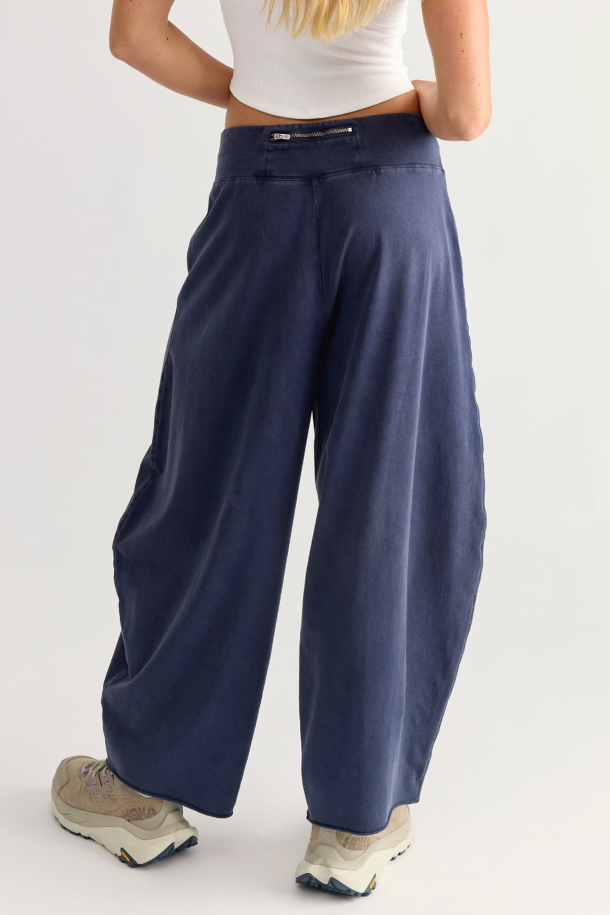 A person shown from the back wearing loose, wide-leg navy pants, a white cropped tank top, and beige sneakers against a plain light gray studio background.