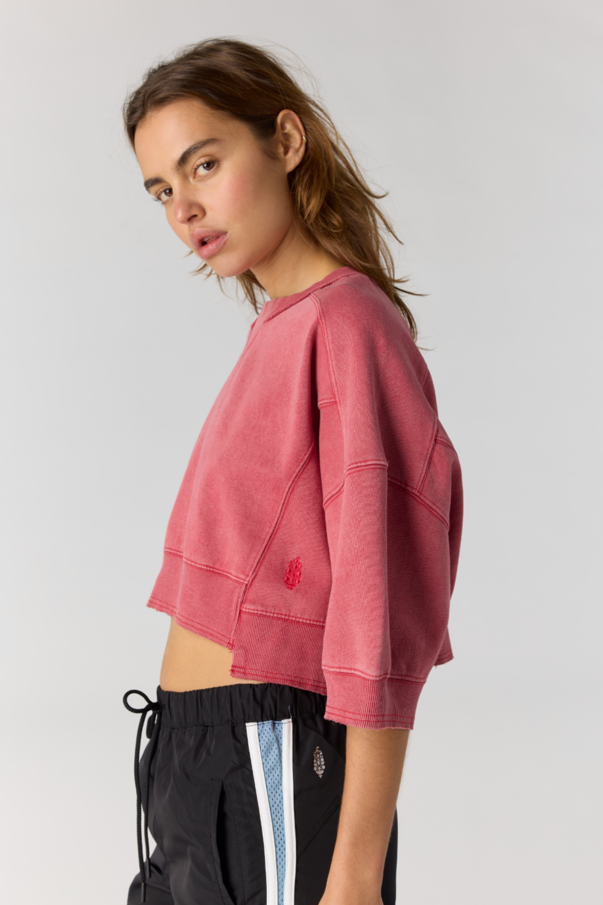 A woman wearing a cropped, faded red sweatshirt and black drawstring pants with a blue and white stripe, photographed against a light gray studio background.