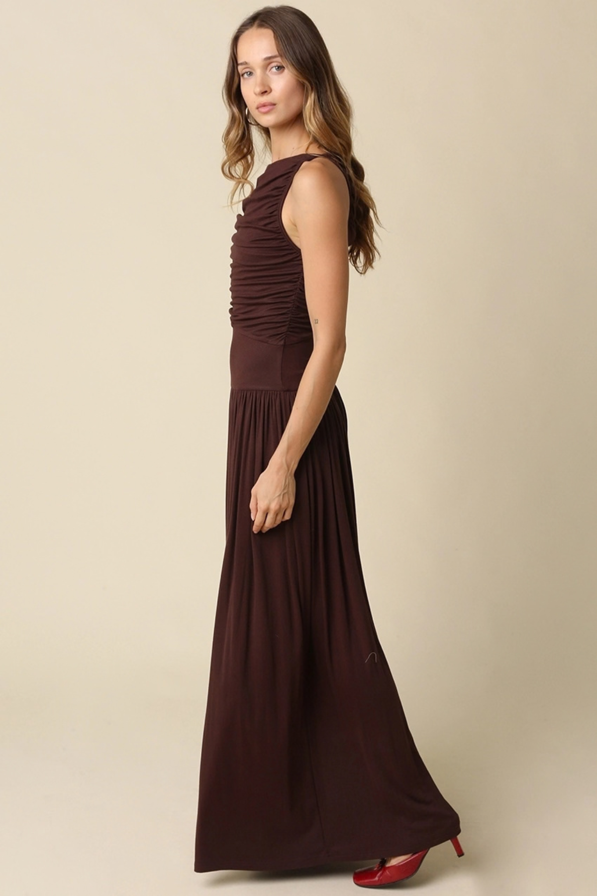 A woman modeling a sleeveless, dark brown maxi dress with a ruched bodice and flowing skirt, paired with red high heels, against a solid beige studio background.