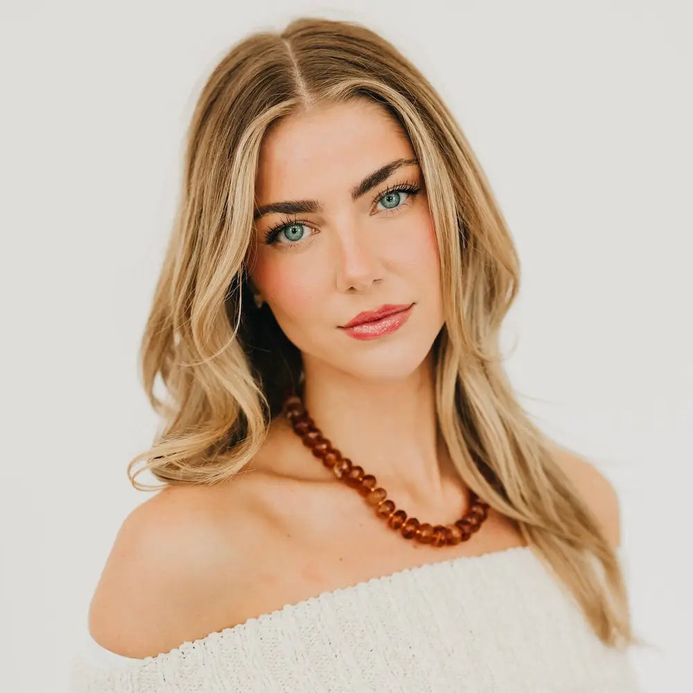 Woman wearing a white off-shoulder top and a brown beaded necklace against a light background