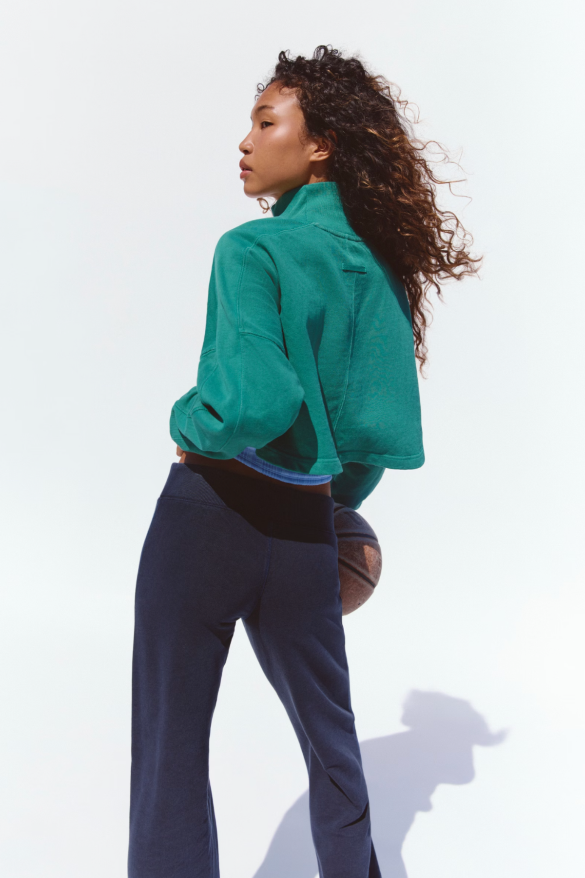 A woman with curly hair is shown from a side-back angle wearing a bright green cropped quarter-zip fleece jacket and navy blue sweatpants, holding a basketball against a clean white studio background.