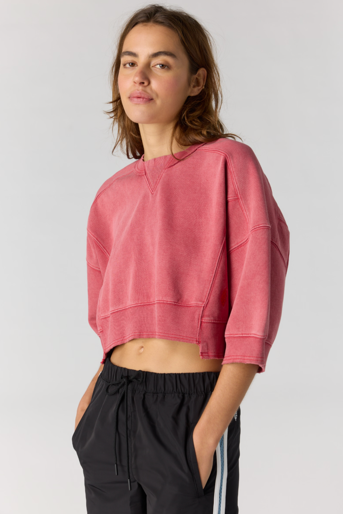 A woman modeling a cropped, three-quarter sleeve red sweatshirt and black athletic pants with white side stripes, photographed against a light gray studio background.