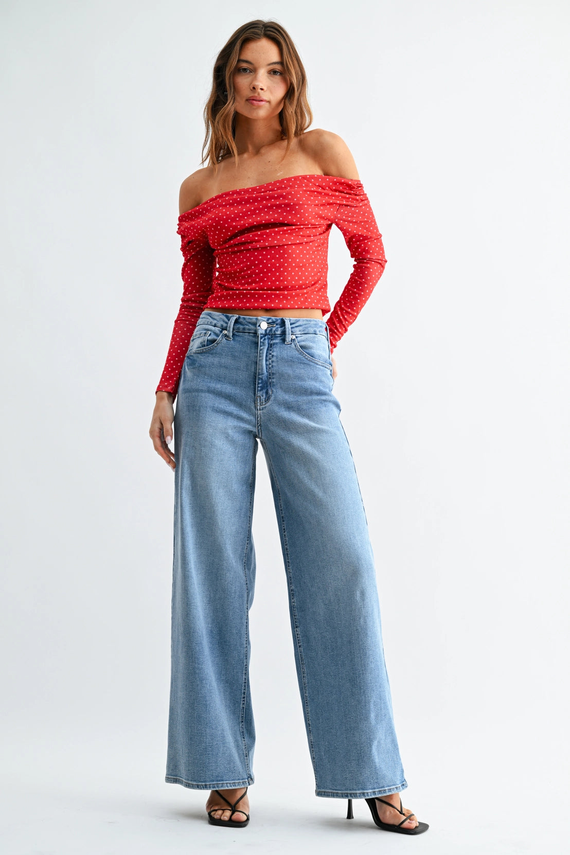 A woman modeling an off-the-shoulder red long-sleeve crop top with a subtle polka dot pattern, paired with high-waisted light blue wide-leg jeans and black strappy heels, against a plain white studio background.