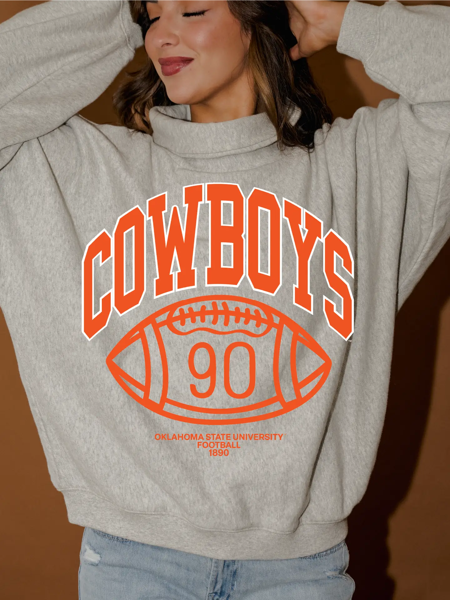 A woman wearing a light heather gray sweatshirt with a large orange and white 'COWBOYS' football graphic on the front, paired with light blue jeans, set against a warm brown background.