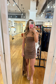 A woman takes a mirror selfie in a clothing boutique wearing a ruched, strapless taupe-brown mini dress. The boutique features a wood floor, clothing racks, and bright overhead lighting.