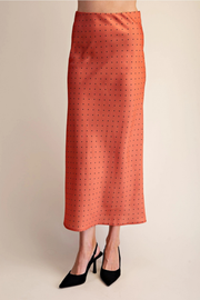 An orange midi skirt with a small black polka dot pattern, shown on a model wearing black pointed-toe heels against a plain beige background.