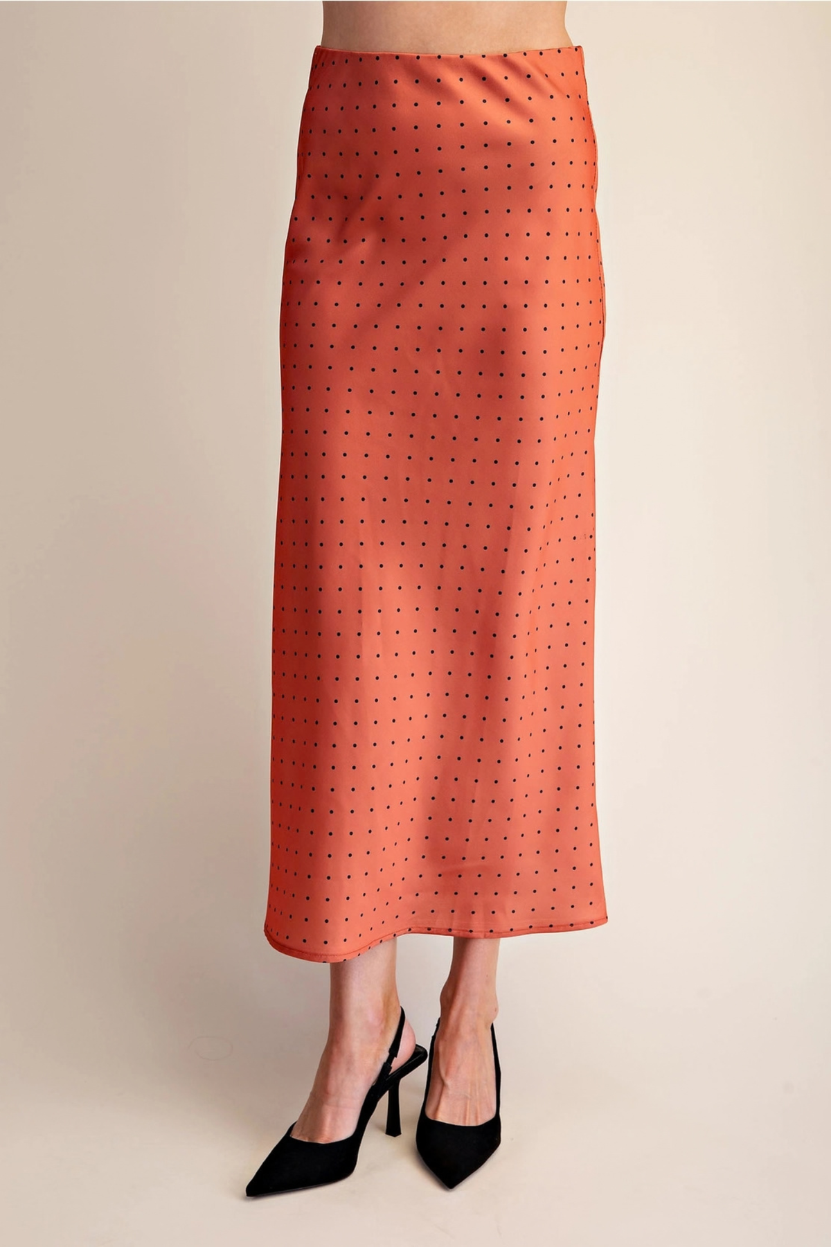 An orange midi skirt with a small black polka dot pattern, shown on a model wearing black pointed-toe heels against a plain beige background.