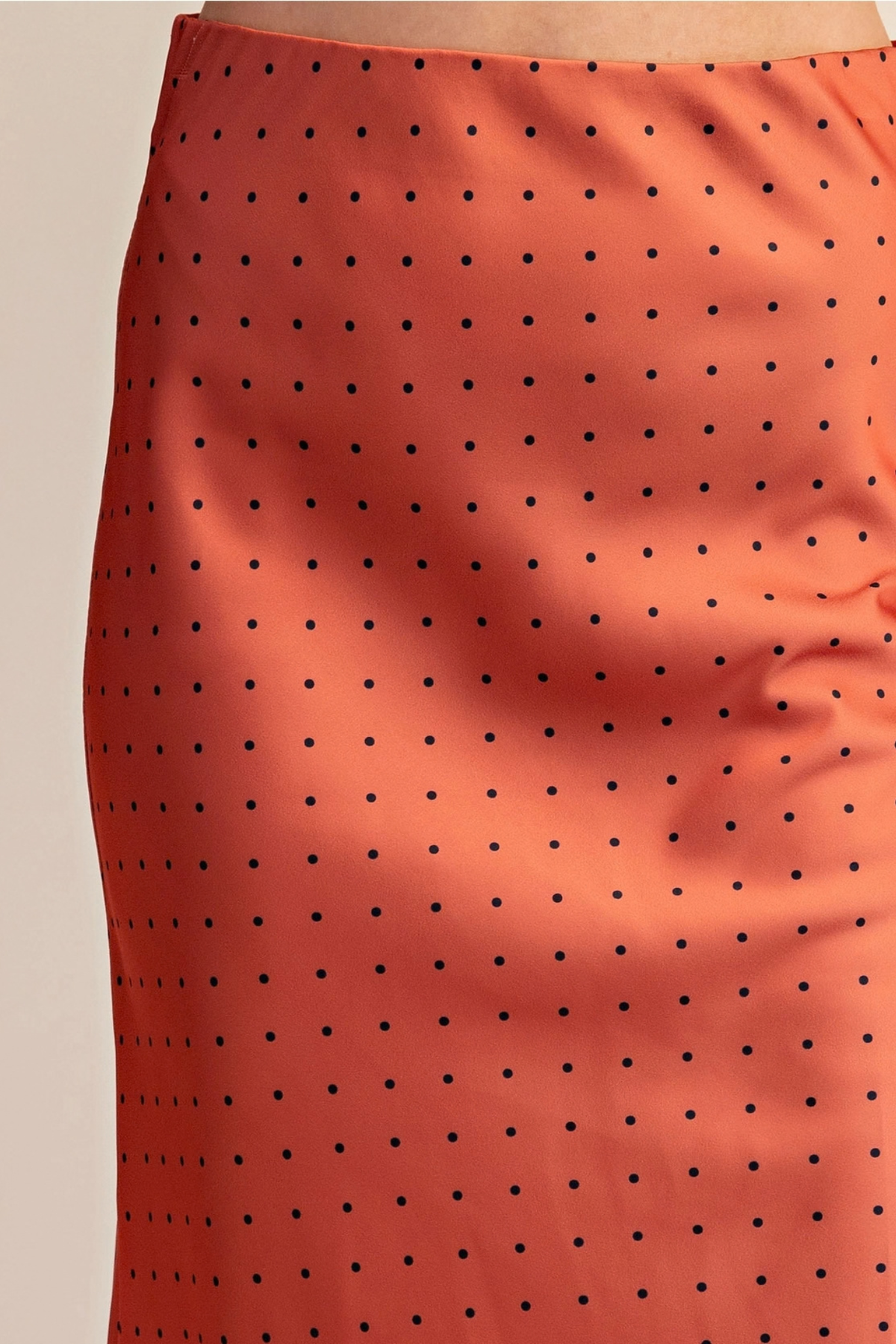 Close-up of a satin-like skirt in a warm coral-orange color with a repeating black polka dot pattern, shown on a model against a light beige background.