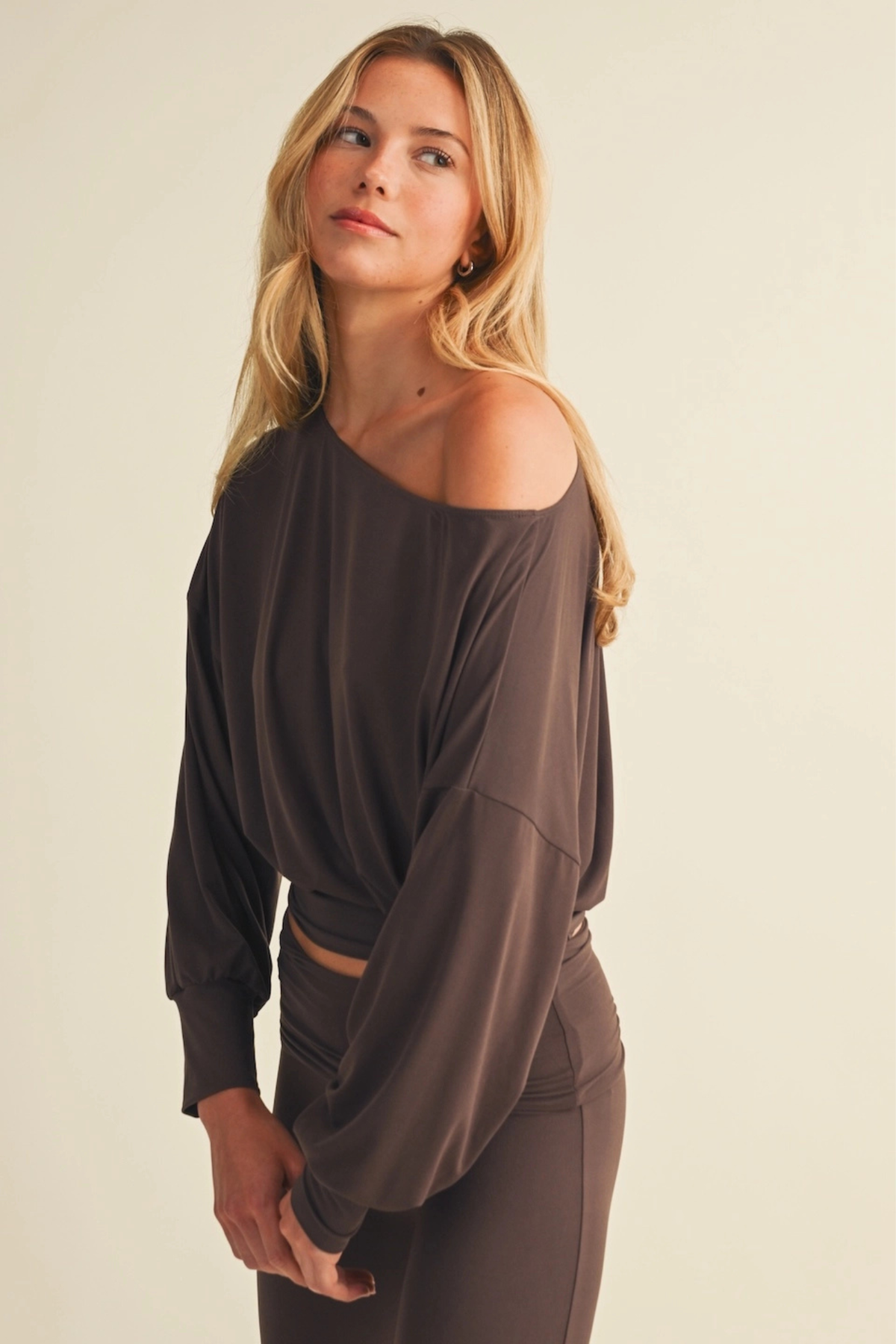 A woman modeling a dark brown off-the-shoulder long-sleeve crop top and matching brown pants against a plain beige studio background. At J.Cole Shoes