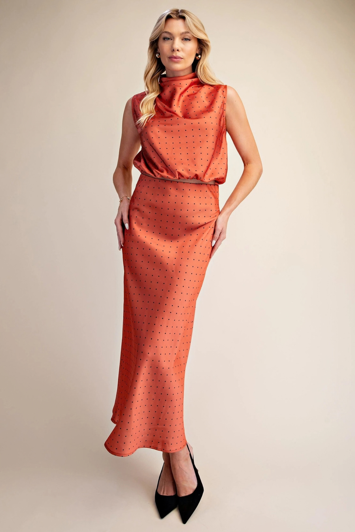 A woman modeling a sleeveless high-neck maxi dress in a warm rust-orange color with a small black polka dot pattern, paired with black pointed-toe heels, against a solid beige studio background.