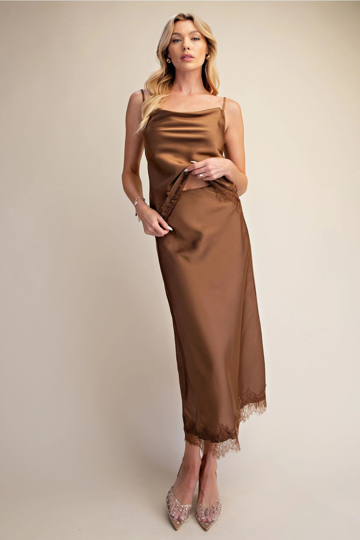 A woman models a satin slip dress in a warm brown color, featuring spaghetti straps, a side tie detail, and a lace-trimmed hem, set against a solid beige studio background.