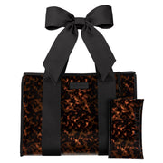 A structured tote bag with a glossy tortoiseshell pattern in brown and black, featuring a large matching black satin bow on the handle and a small matching zip pouch placed in front. The bag is photographed on a clean white background.