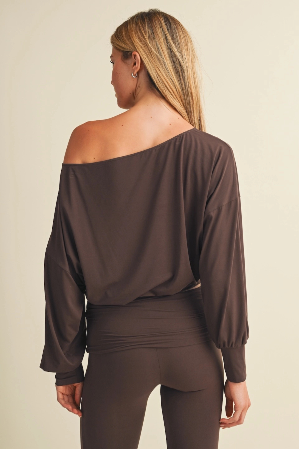 A woman shown from the back wearing a dark brown off-the-shoulder long-sleeve top and matching dark brown leggings against a plain light beige studio background.
