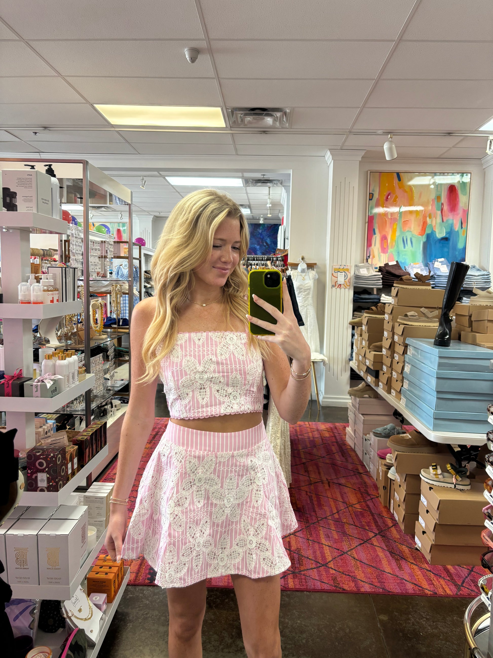A woman takes a mirror selfie in a retail store while wearing a light pink and white lace crop top and matching mini skirt.