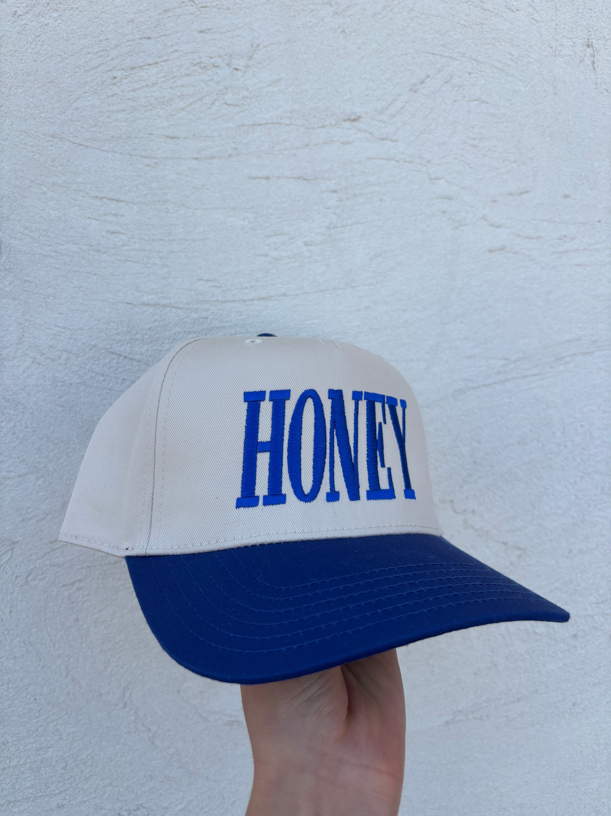 A white and navy blue trucker-style cap with the word 'HONEY' embroidered in blue on the front, held by a hand against a textured white wall.