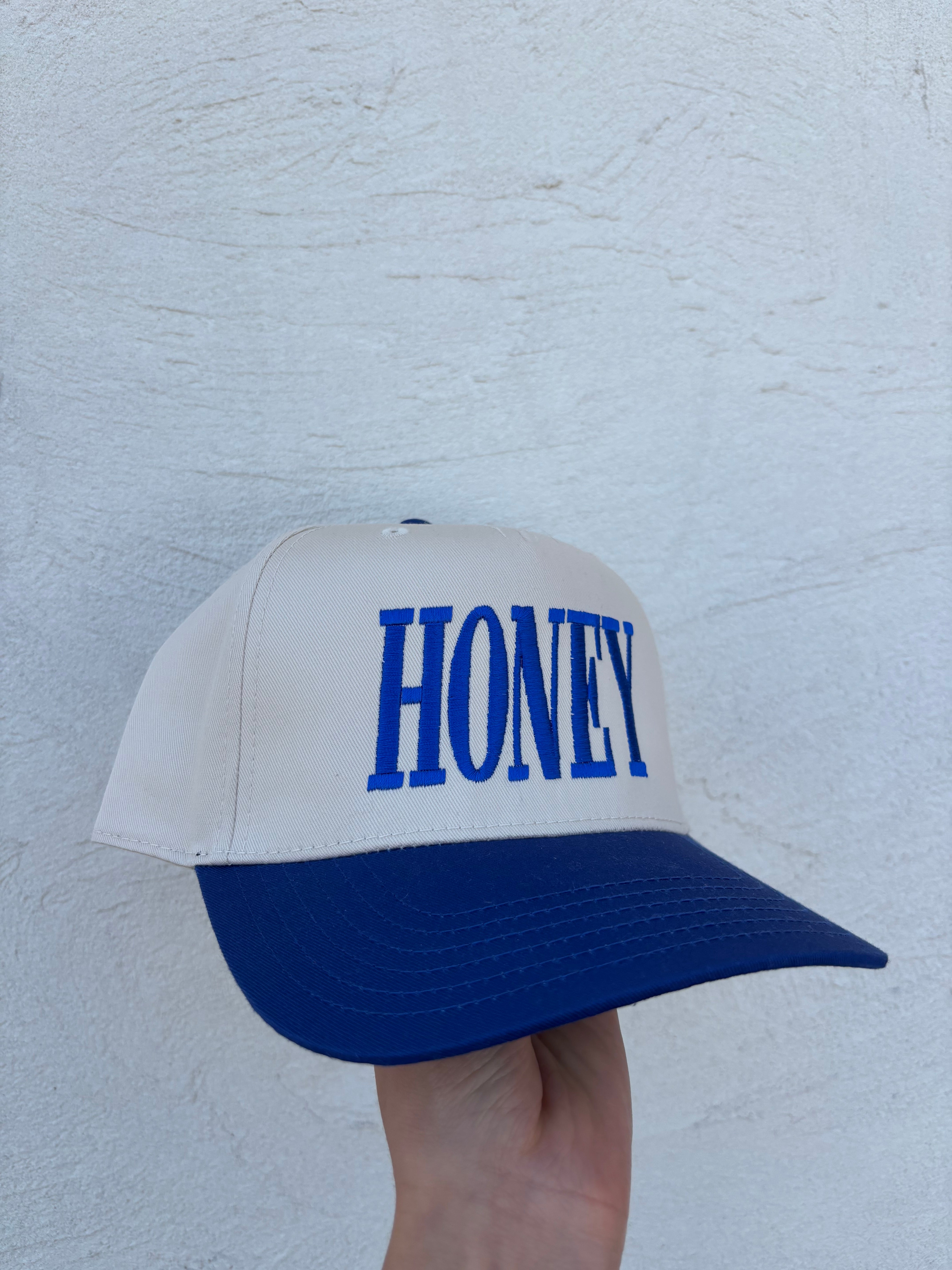A white and navy blue trucker-style cap with the word 'HONEY' embroidered in blue on the front, held by a hand against a textured white wall.