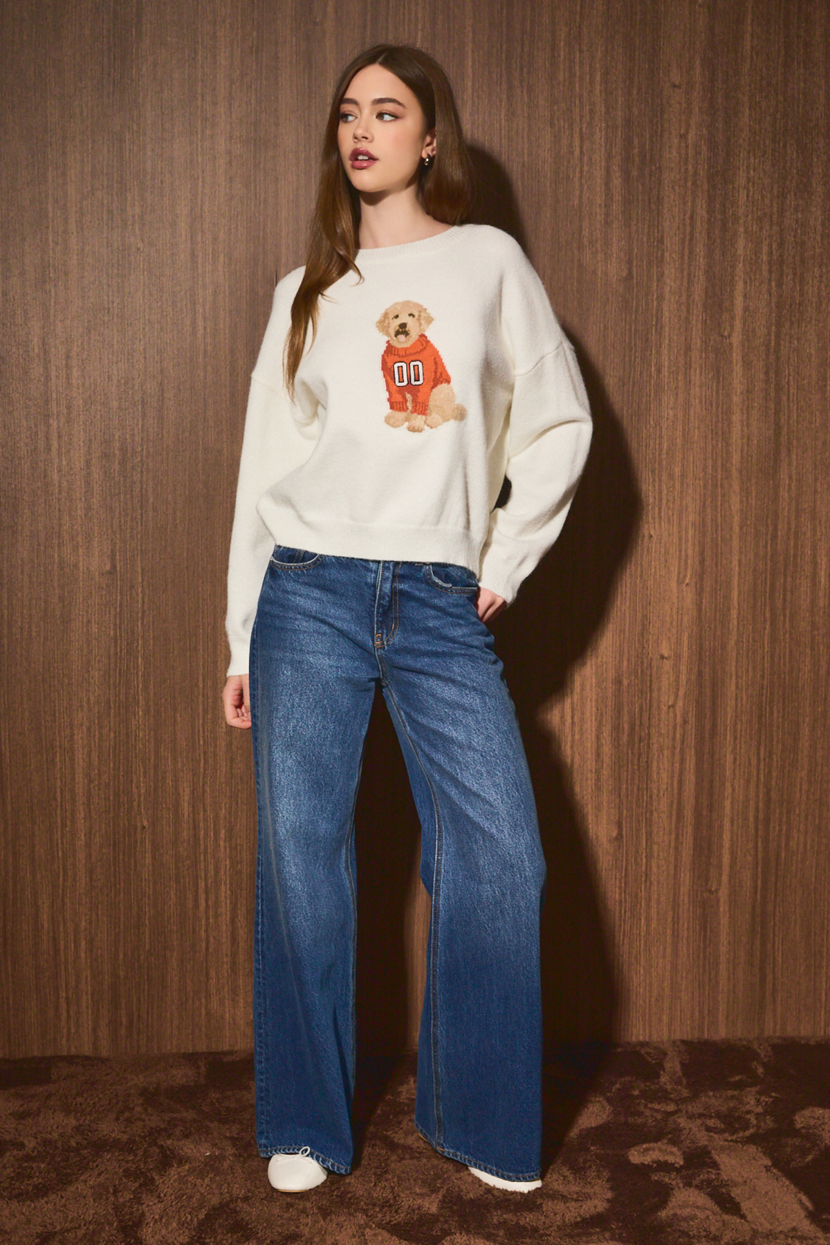 A woman stands against a wood-paneled wall wearing a cream sweatshirt with a dog graphic and blue wide-leg jeans.