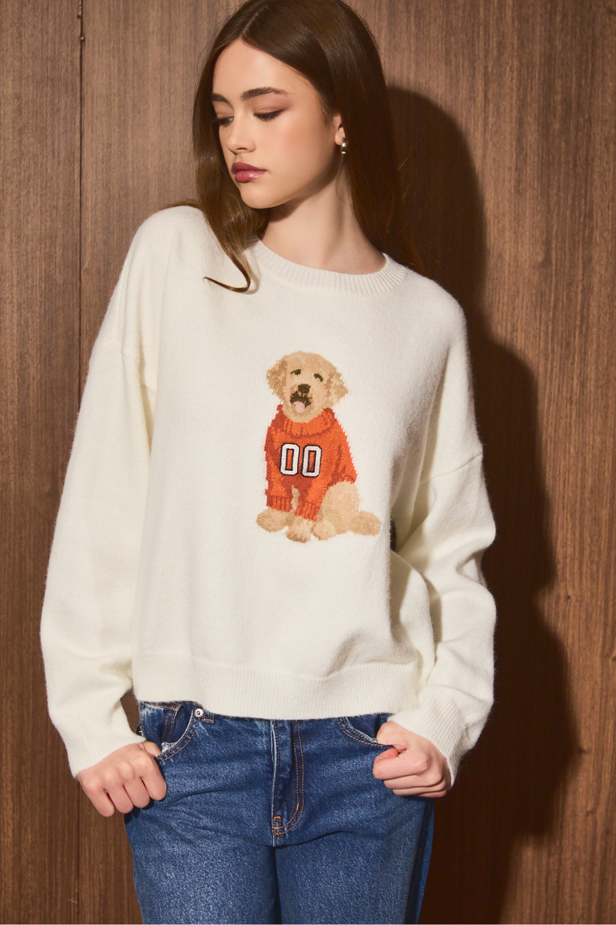 A woman wearing a cream-colored crewneck sweater with a graphic of a golden retriever dog in an orange sports jersey, paired with blue jeans, standing against a dark wood-paneled wall.