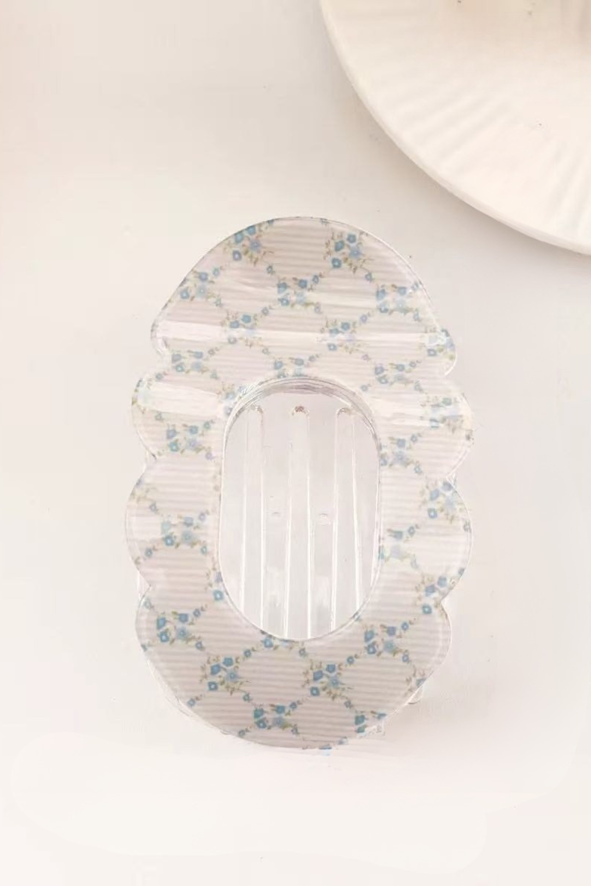A clear plastic Hair Clip with a scalloped edge and a delicate blue and white floral pattern, shown on a light off-white background.