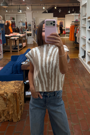 A woman takes a mirror selfie in a clothing boutique wearing a sleeveless knit top with a beige and white tiger-like stripe pattern, paired with blue jeans.