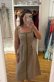 A woman takes a mirror selfie in a clothing boutique wearing a sleeveless midi dress with vertical brown and beige stripes and olive green trim. The background shows a clothing rack with various garments and white hats on a shelf.