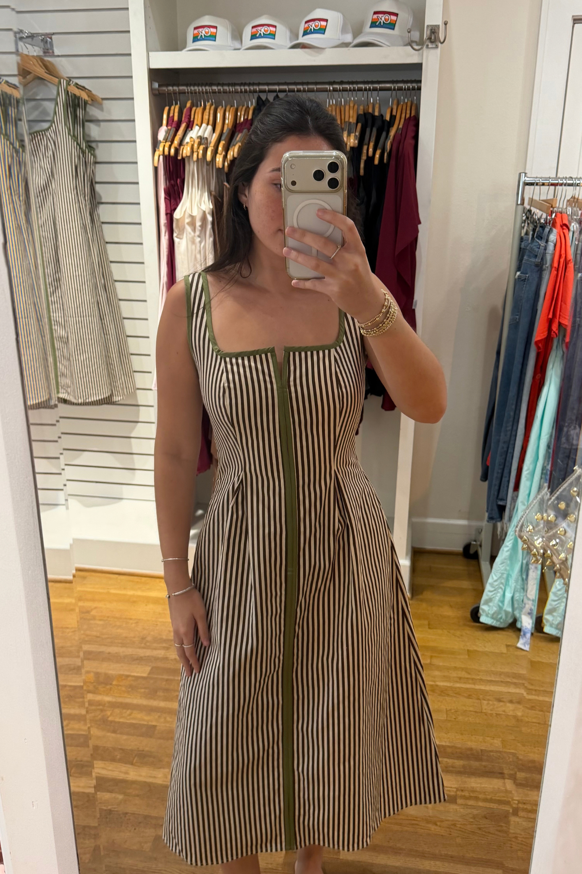 A woman takes a mirror selfie in a clothing boutique wearing a sleeveless midi dress with vertical brown and beige stripes and olive green trim. The background shows a clothing rack with various garments and white hats on a shelf.