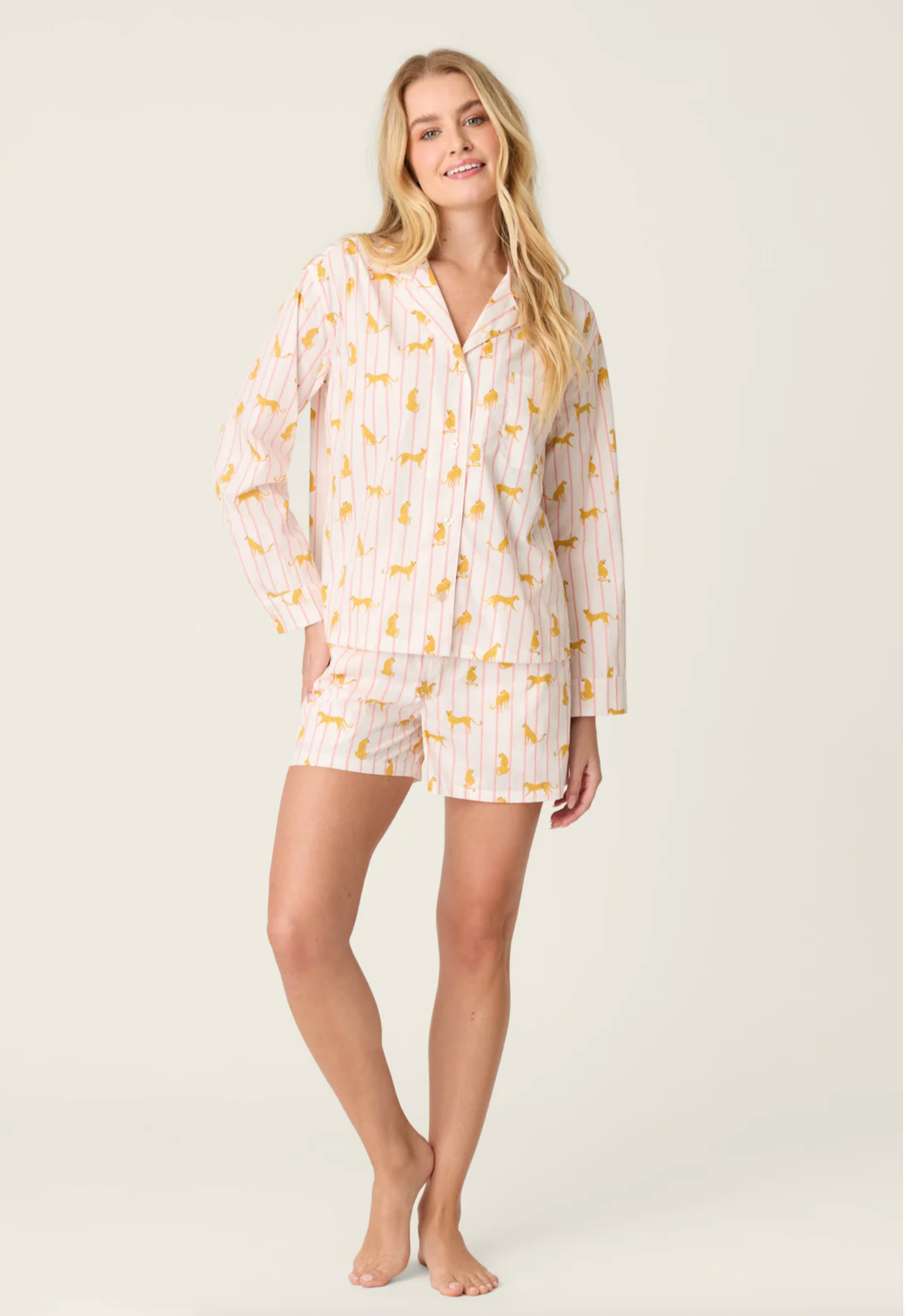 A woman modeling a two-piece pajama set consisting of a long-sleeve button-up shirt and matching shorts, featuring a white base with thin vertical yellow stripes and a scattered yellow floral pattern, set against a plain light beige studio background.