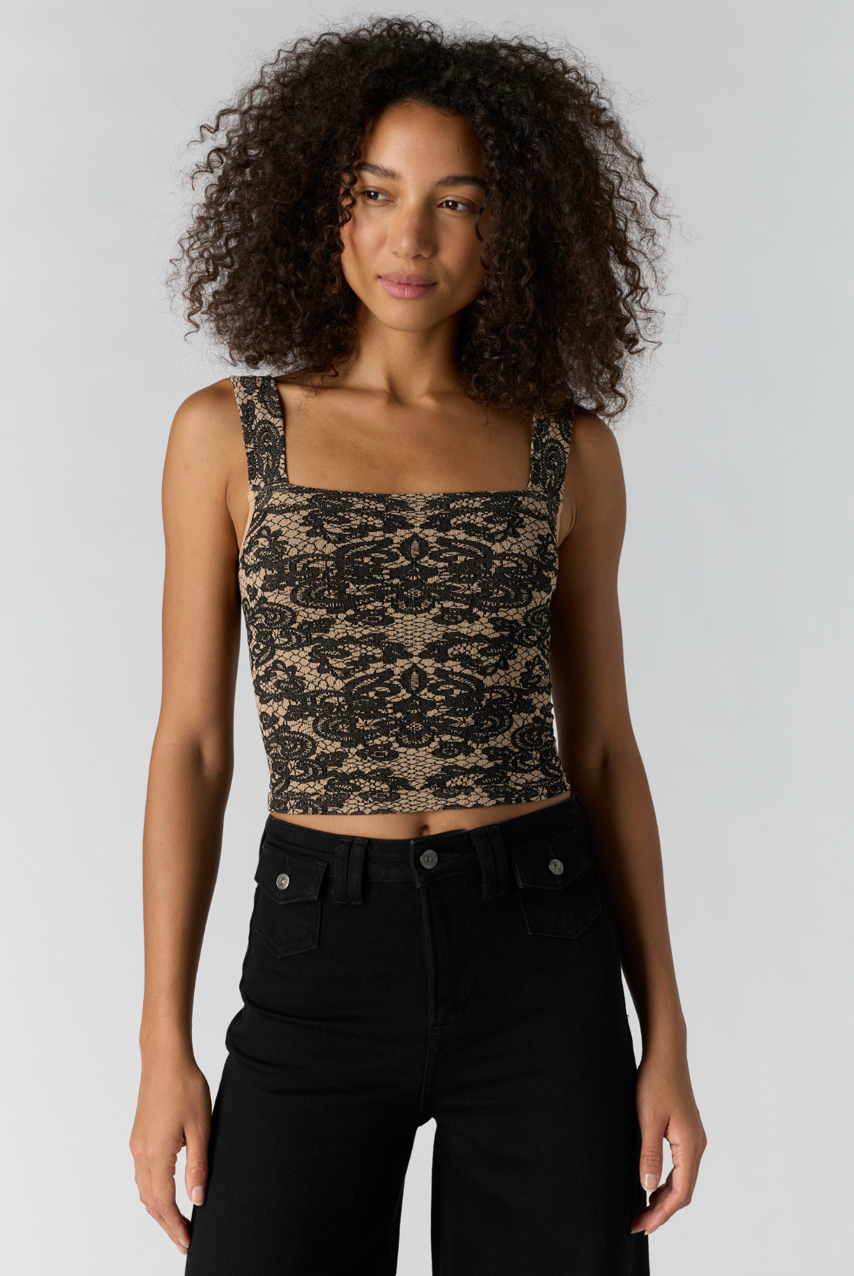 A woman with curly dark hair wearing a black and beige patterned lace crop top and black jeans, standing against a light gray studio background.