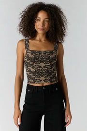 A woman with curly dark hair wearing a black and beige patterned lace crop top and black jeans, standing against a light gray studio background.
