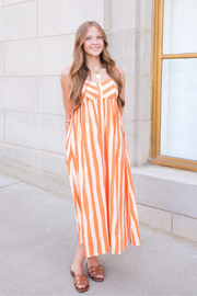 A woman wearing a sleeveless orange and white vertically striped maxi dress and brown strappy sandals, standing outdoors against a light stone building wall.